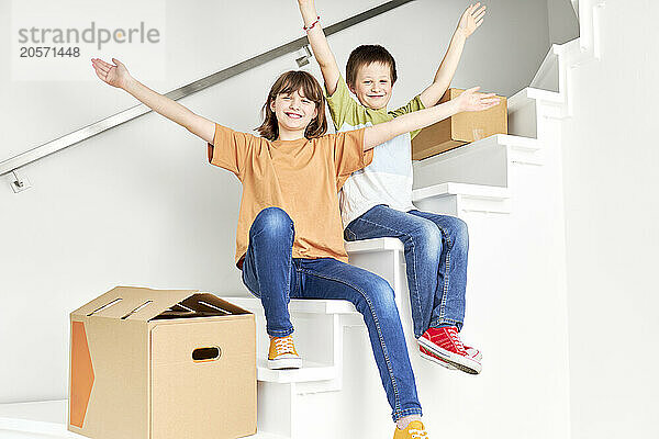 Happy boy and girl sitting with arms outstretched amidst boxes on staircase