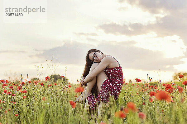 Young woman with eyeglasses hugging legs and sitting on stool in poppy field