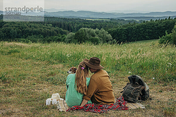 Boyfriend and girlfriend with dog sitting on blanket in meadow at mountain