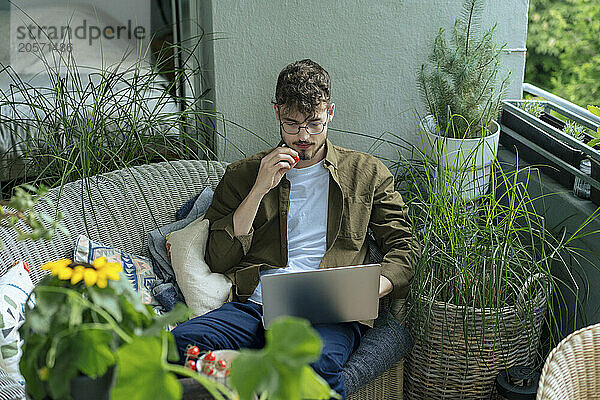 Handsome young man eating cherry tomato and using laptop sitting in balcony