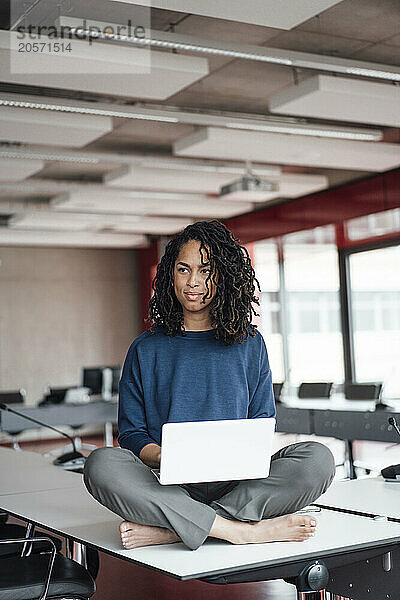 Thoughtful businesswoman sitting cross-legged with laptop on conference table at office