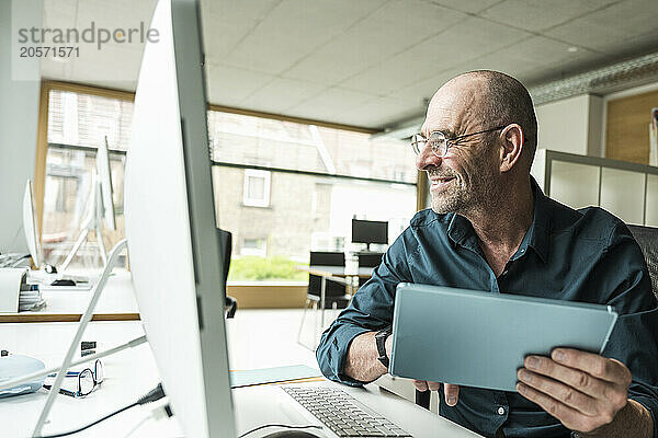 Smiling mature businessman with tablet PC looking at computer monitor on desk in office