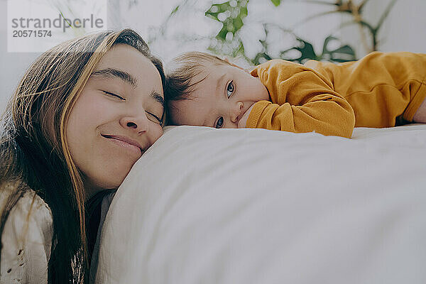 Affectionate mother leaning on bed with baby boy lying on front in bed at home