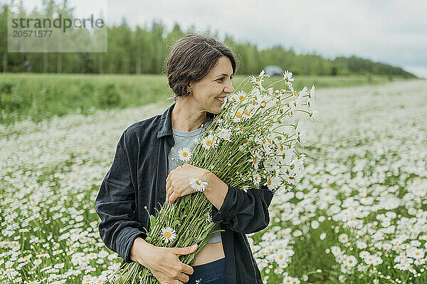 Happy mature woman standing with bouquet of daisy flowers on field