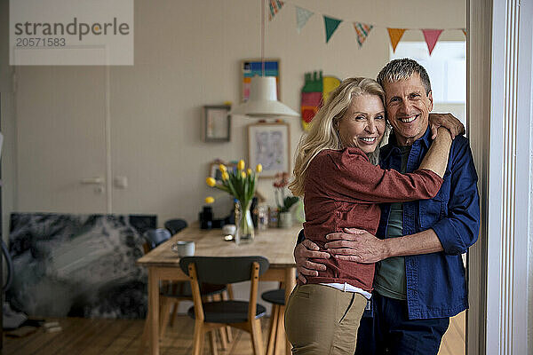 Smiling senior woman with arm around man leaning on wall at home