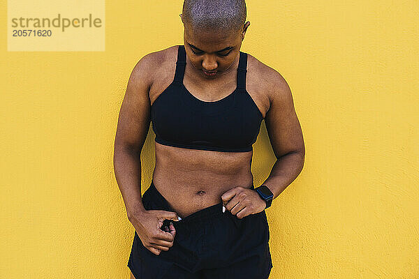 Bald woman wearing shorts and standing in front of yellow wall