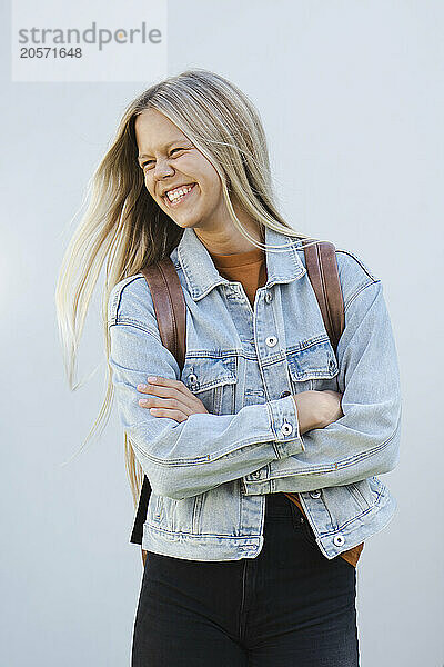 Cheerful long hair teenager in denim jacket with arms crossed standing in front of wall