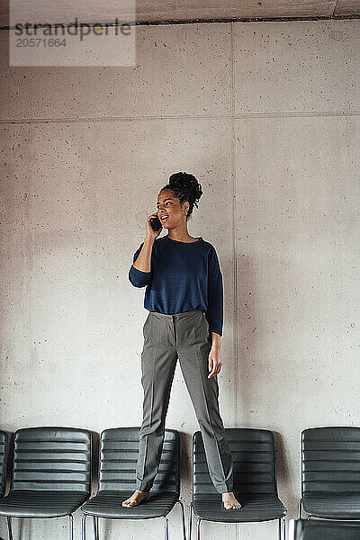 Young businesswoman standing on chairs talking through mobile phone in front of wall at office lobby