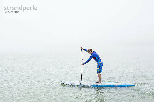 Man wearing blue wetsuit paddleboarding on lake in foggy weather