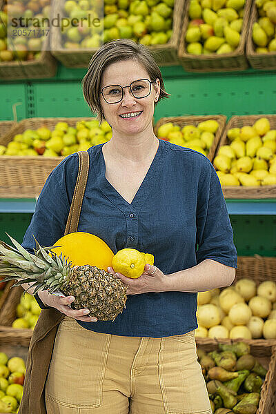 Smiling mature woman in eyeglasses carrying fruits and standing in supermarket