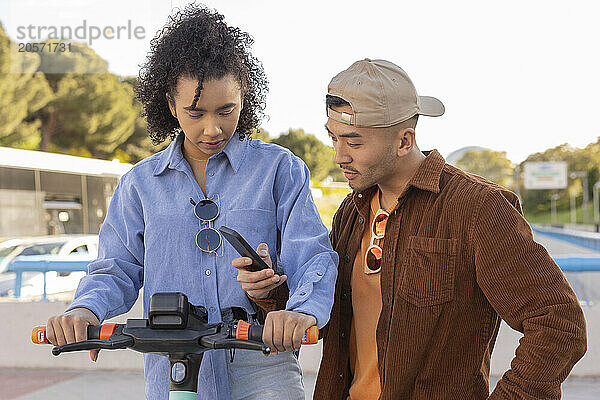 Young man unlocking push scooter through smart phone with girlfriend at park