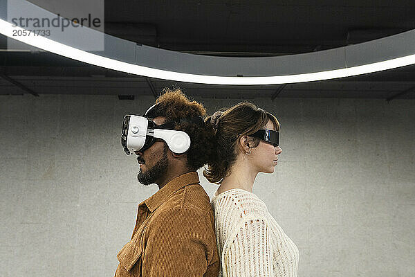 Multiracial couple wearing virtual reality simulators standing back to back in parking garage