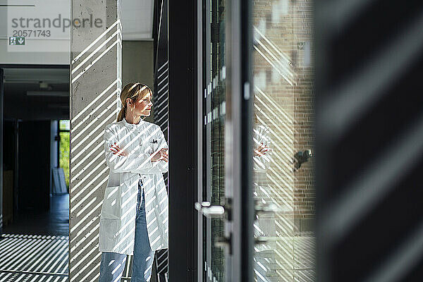 Confident female doctor with arms crossed looking out through window in hospital