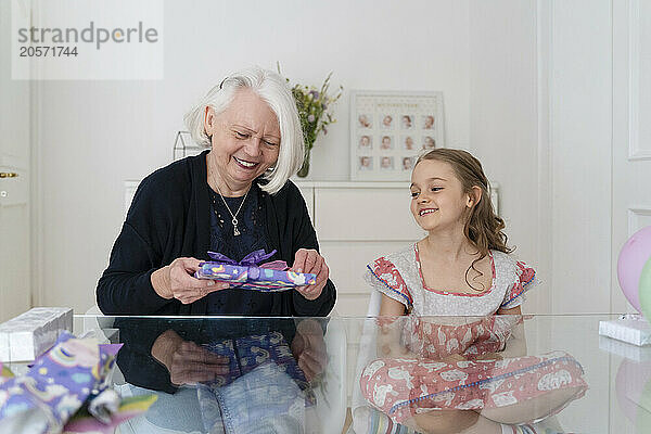 Smiling senior woman with gift sitting by granddaughter at home