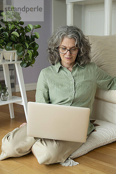 Gray hair businesswoman working on laptop and sitting on cushion at home