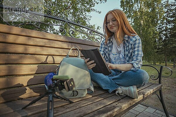 Teenage freelancer using tablet PC sitting cross-legged on bench and vlogging through smart phone at public park