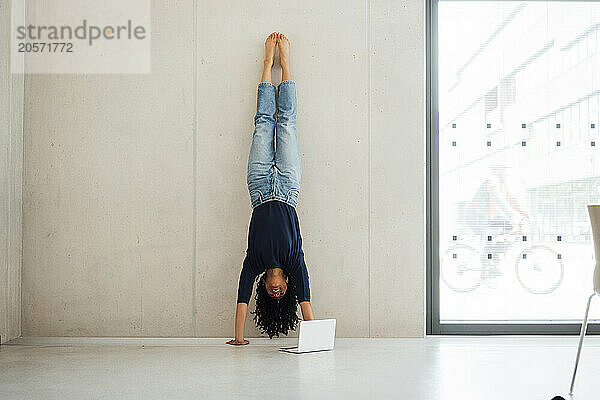 Young businesswoman leaning on wall doing handstand near laptop at office lobby