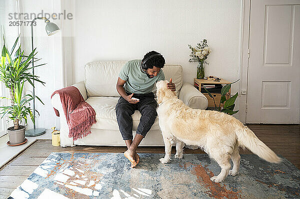 Young man stroking dog sitting on sofa in living room at home