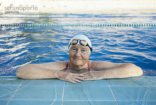 Smiling senior woman in swimming cap and googles resting on edge of pool