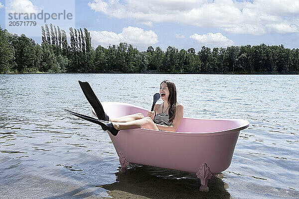 Cheerful girl with diving flipper holding back brush and sitting inside pink bathtub in lake