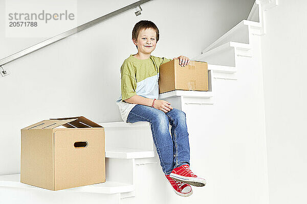 Smiling boy sitting with boxes on staircase of new house