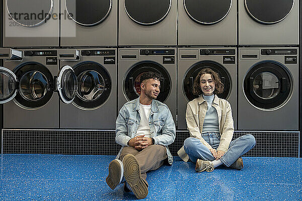 Smiling young man and woman sitting together leaning on washing machines in laundromat