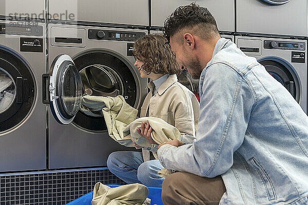 Smiling young couple removing sheet from washing machine at laundromat