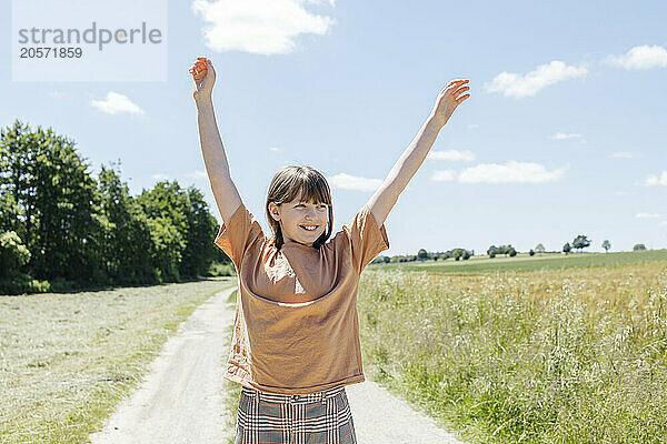 Smiling girl with arms raised standing on footpath between meadow