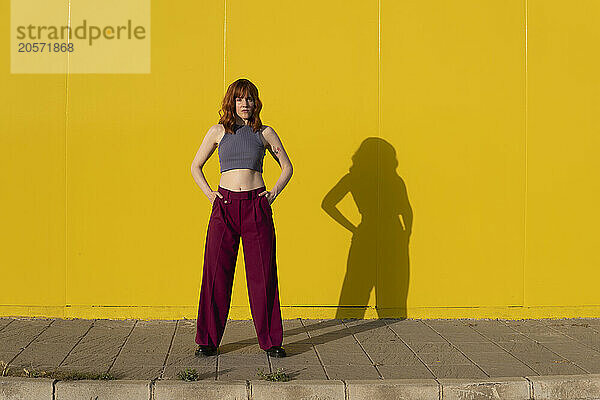 Confident redhead woman standing on footpath in front of yellow wall