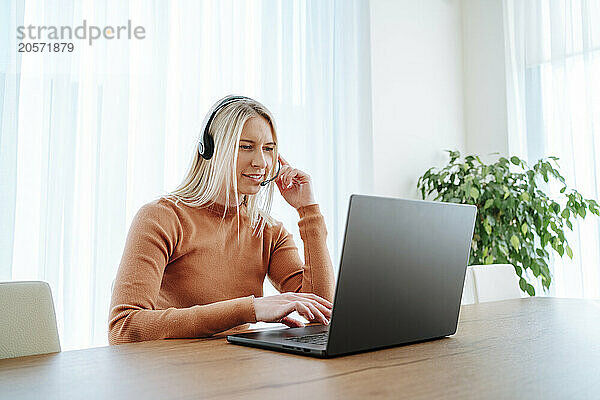 Young blond businesswoman with headset using laptop at table working from home