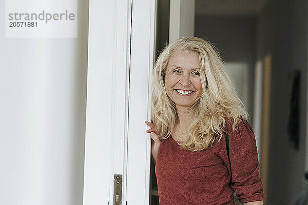 Smiling blond senior woman leaning on doorway at home