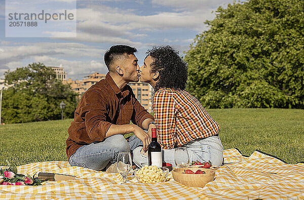 Boyfriend and girlfriend kissing and sitting on picnic blanket in public park