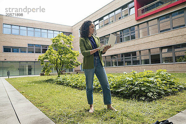 Confident young businesswoman using laptop standing on grass at office park