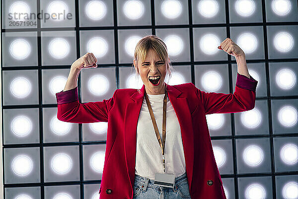 Playful blond businesswoman flexing muscles in front of illuminated wall at office