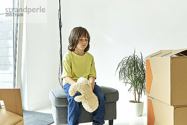 Sad boy with stuffed toy sitting on ottoman stool in new house