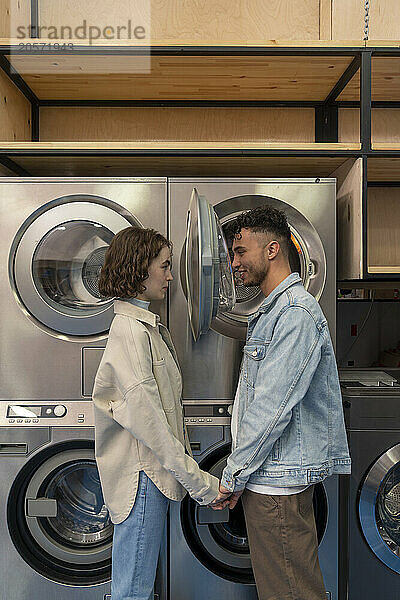 Young couple holding hands and looking through washing machine glass at laundromat