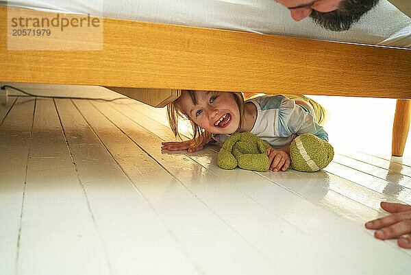 Cheerful girl hiding under bed at home