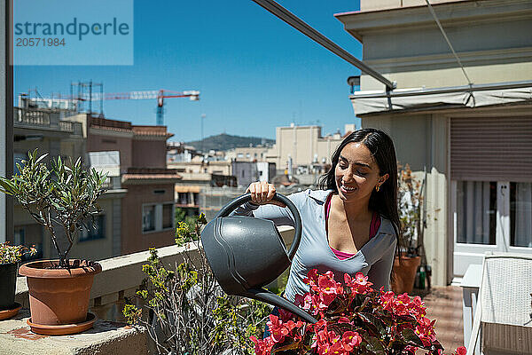 Smiling woman watering fresh red flowers at rooftop on sunny day