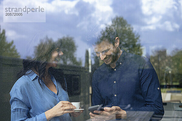 Happy businessman using smart phone with colleague holding coffee cup seen through window