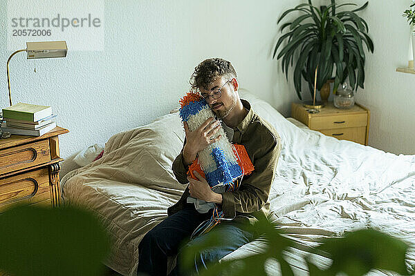 Young man with eyes closed hugging rocketship shaped pinata sitting on bed at home