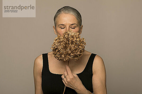 Senior woman with eyes closed holding withered flower and covering face against brown background