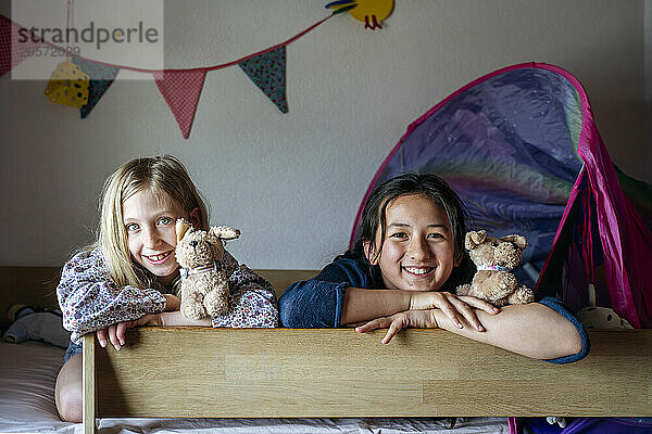 Smiling girls with stuffed toy animals leaning on bunk bed at home