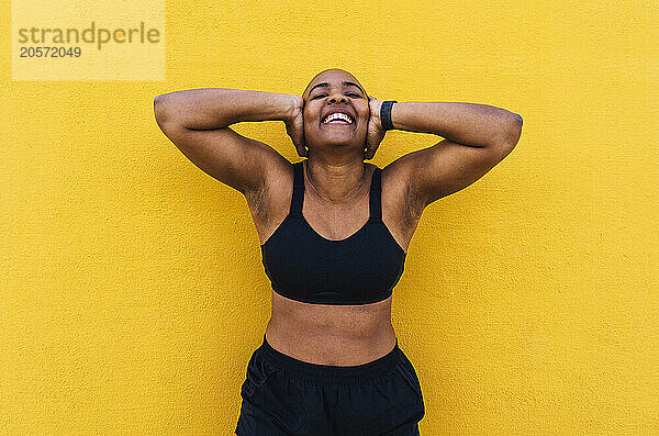 Cheerful bald woman covering ears with hands in front of yellow wall