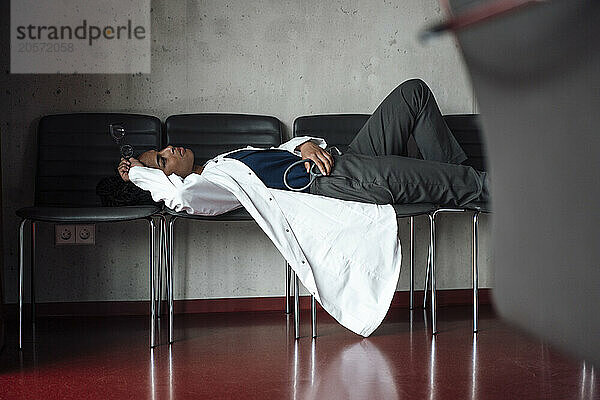 Tired young female doctor lying on chairs at hospital lobby