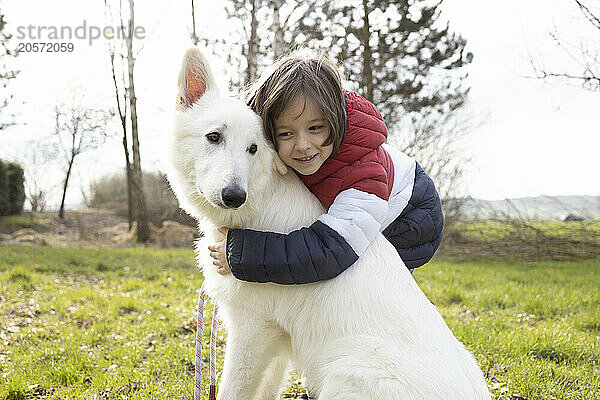 Cute boy embracing White Swiss Shepherd Dog on grass in meadow