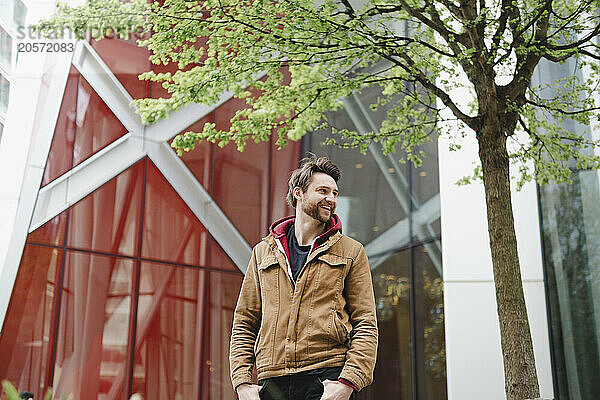 Happy man with hands in pockets standing near tree