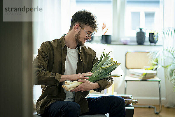 Smiling young man sitting with fresh leek vegetable at home