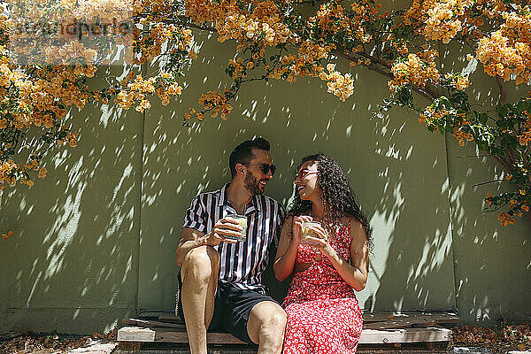 Happy couple enjoying iced drinks sitting in shade near wall