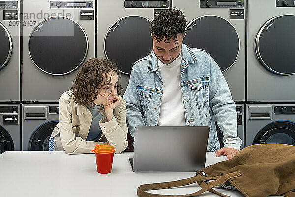 Young couple using laptop on table in front of washing machines waiting at laundromat