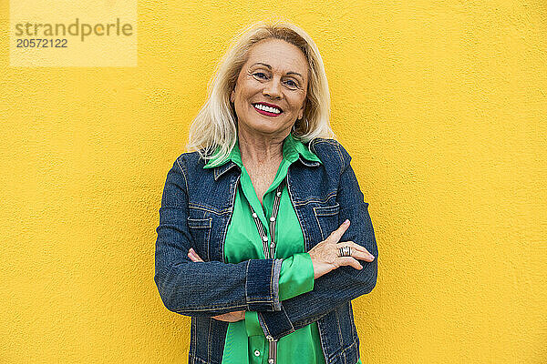 Happy senior woman with arms crossed in front of yellow wall